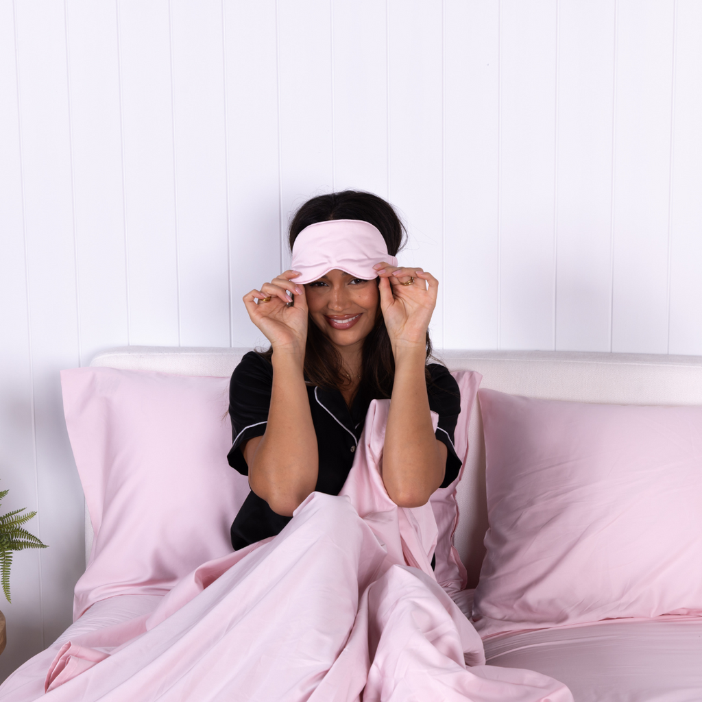 Smiling woman in black pyjamas adjusting blush pink sleep mask while sitting in bed with The Lad Collective bamboo cotton blend blush bedding set, highlighting comfort and relaxation.