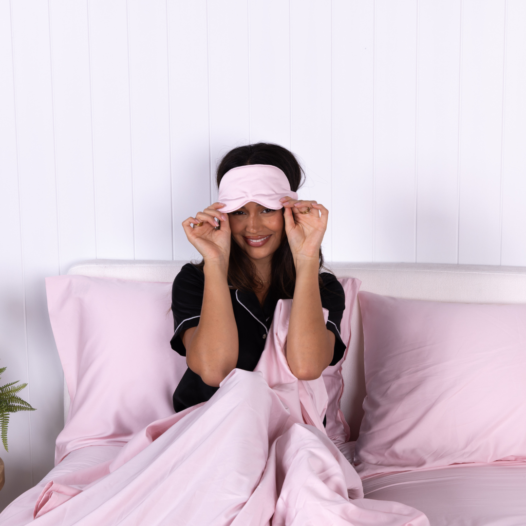 Smiling woman in black pyjamas adjusting blush pink sleep mask while sitting in bed with The Lad Collective bamboo cotton blend blush bedding set, highlighting comfort and relaxation.