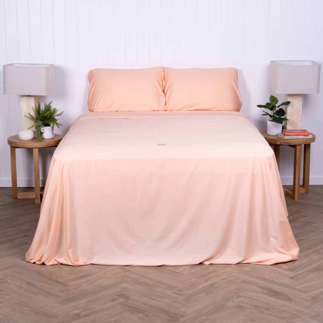 Peach bedding set neatly styled on a double bed with matching pillowcases – soft, breathable sheets featured in a bright, modern bedroom with timber bedside tables, neutral lamps and lush greenery.