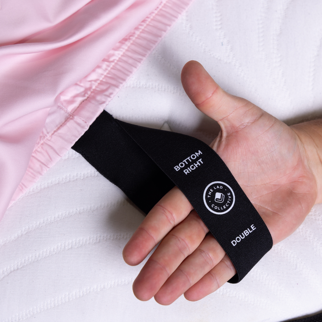 Hand holding labeled corner tag of The Lad Collective fitted sheet in soft pink, showing “Bottom Right” and “Double” size indicator for easy bed-making.