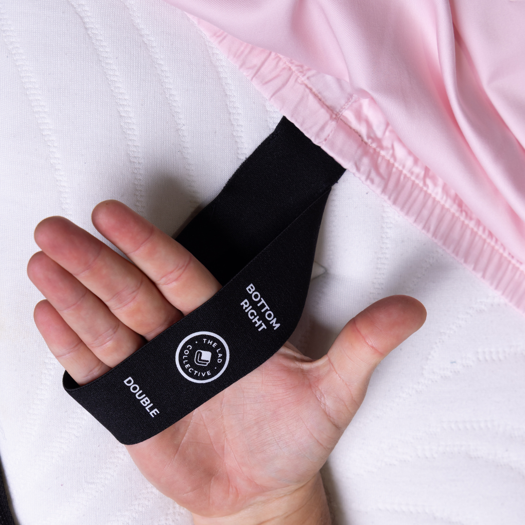 Hand holding labeled corner tag of The Lad Collective fitted sheet in soft pink, showing “Bottom Right” and “Double” size indicator for easy bed-making.