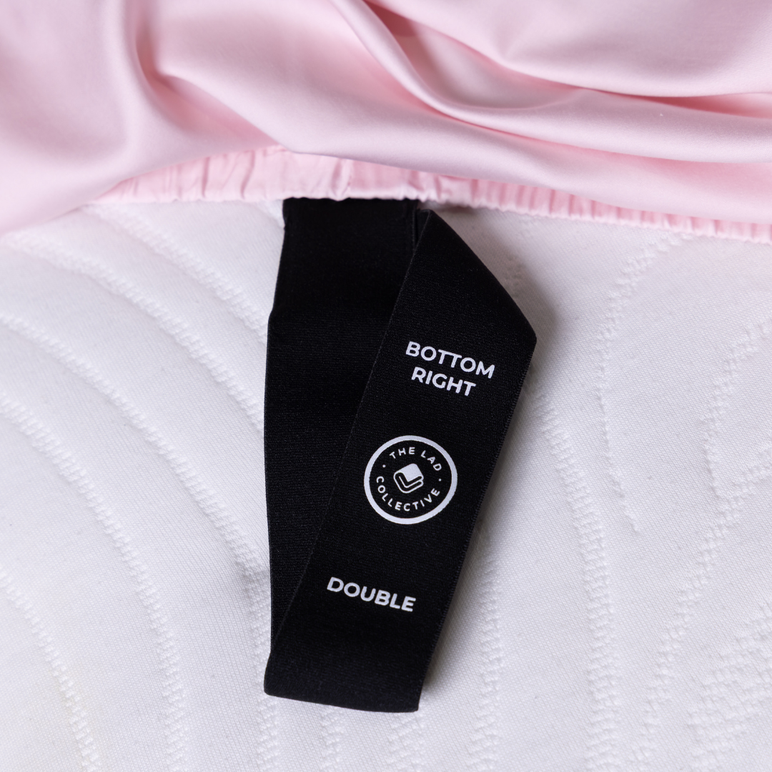 Labeled corner tag of The Lad Collective fitted sheet in soft pink, showing “Bottom Right” and “Double” size indicator for easy bed-making.
