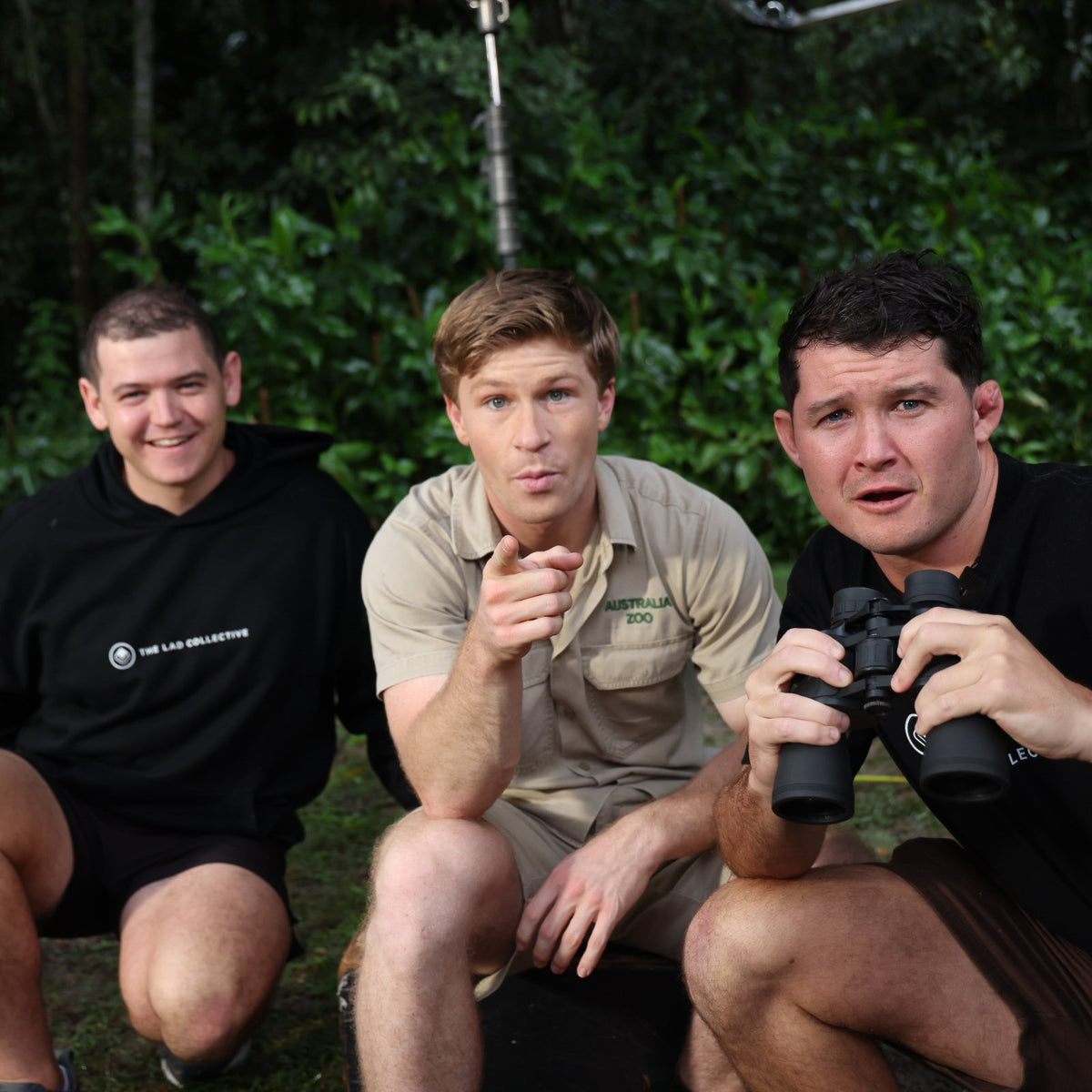 Robert Irwin crouching outdoors with two men, one holding binoculars