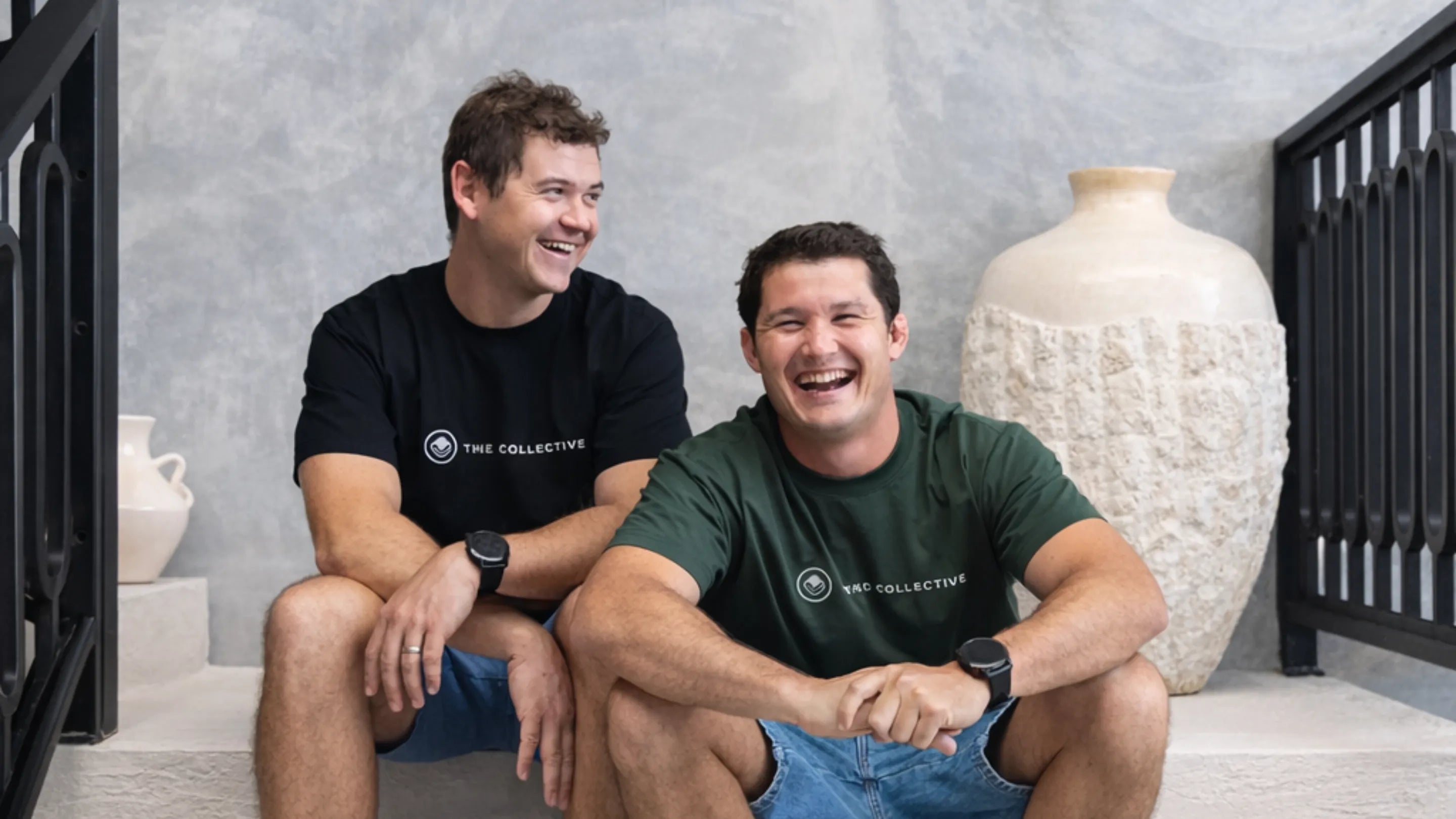 Two men sitting on a staircase wearing branded t-shirts with decorative vases in the background.