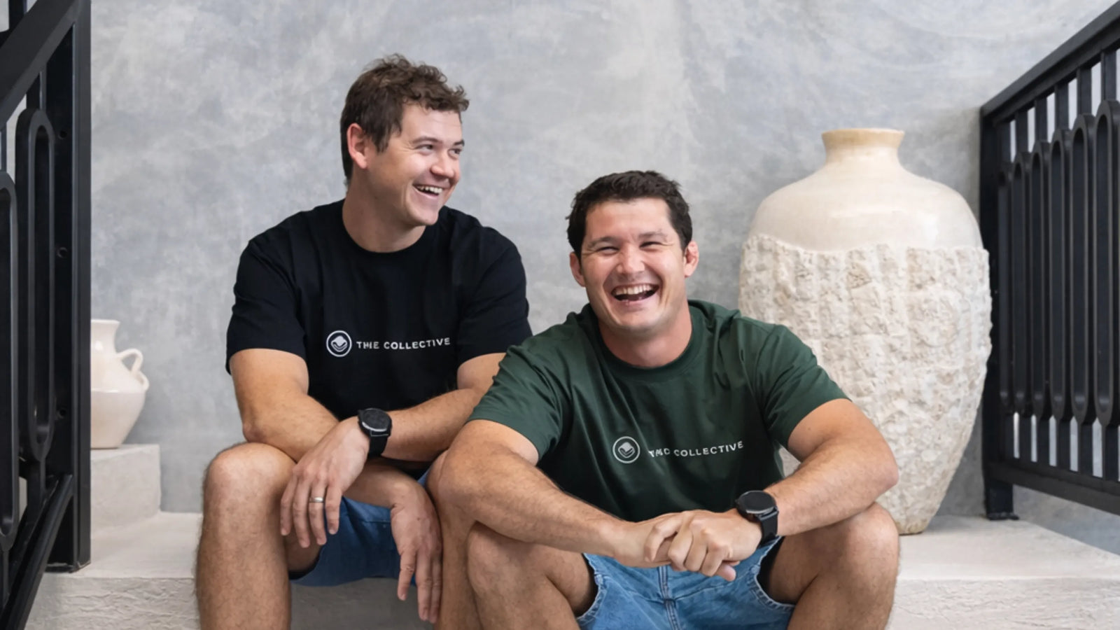 Two men sitting on a staircase wearing branded t-shirts with decorative vases in the background.