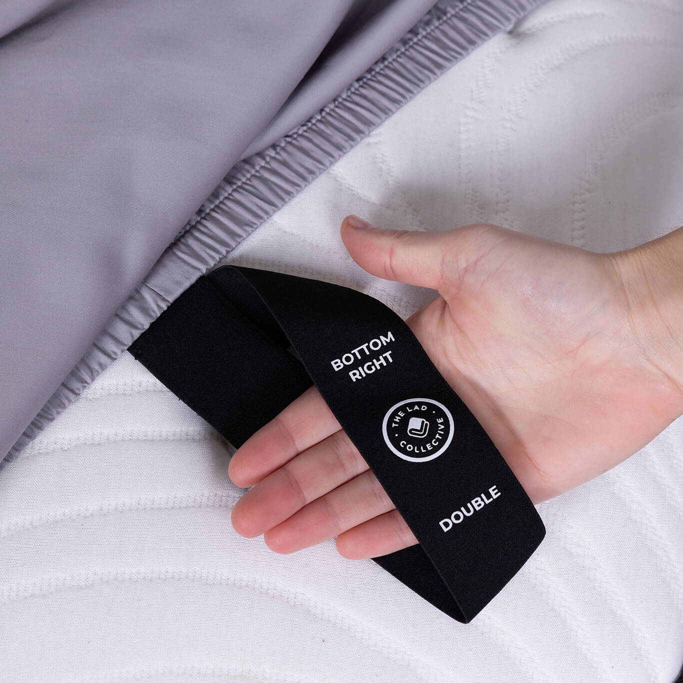 Hand holding a fitted sheet black label with text on top of a mattress.