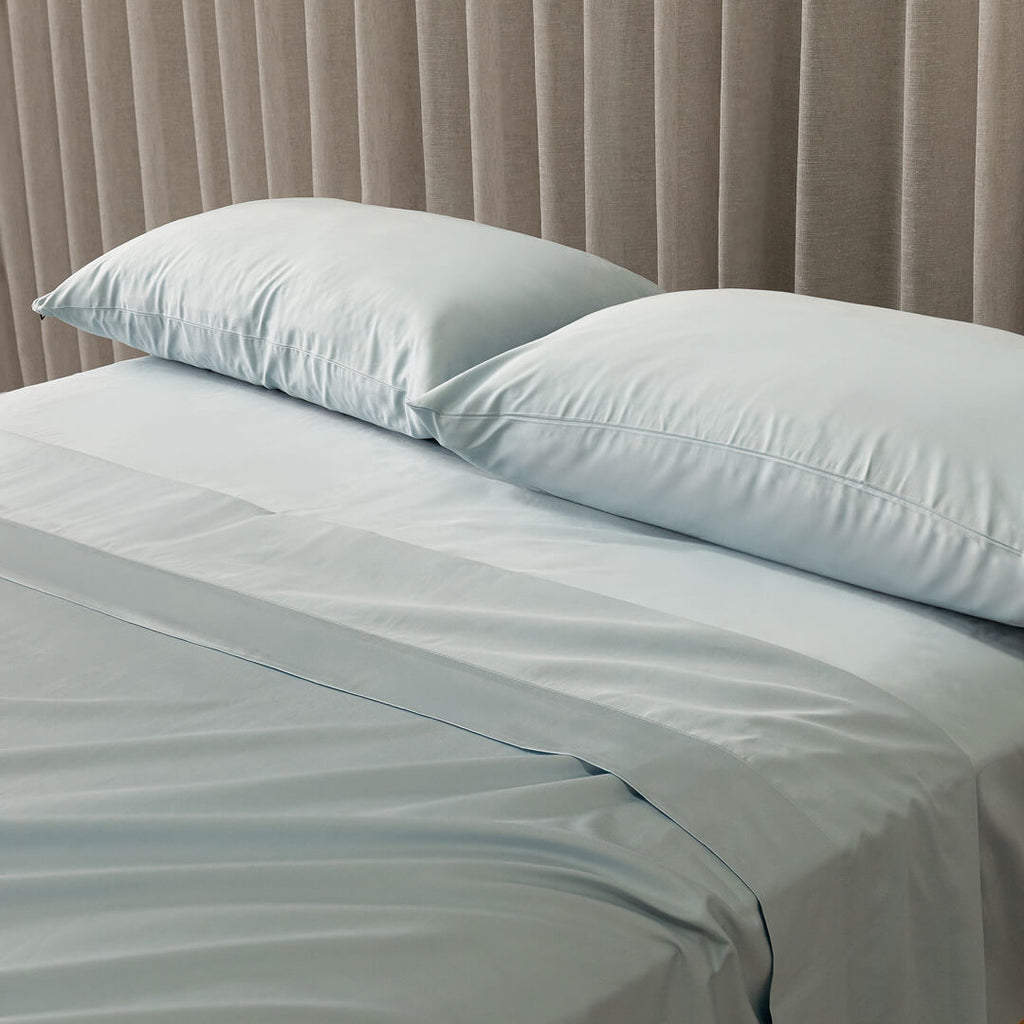 Light blue bedding set with two pillows on a bed against a beige curtain background.