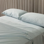Light blue bedding set with two pillows on a bed against a beige curtain background.