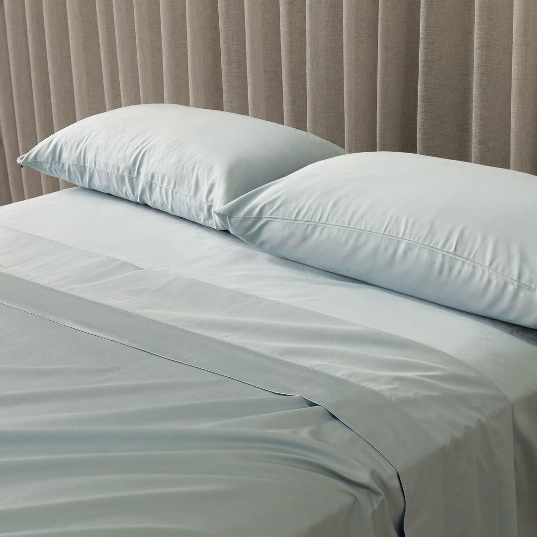 Light blue bedding set with two pillows on a bed against a beige curtain background.