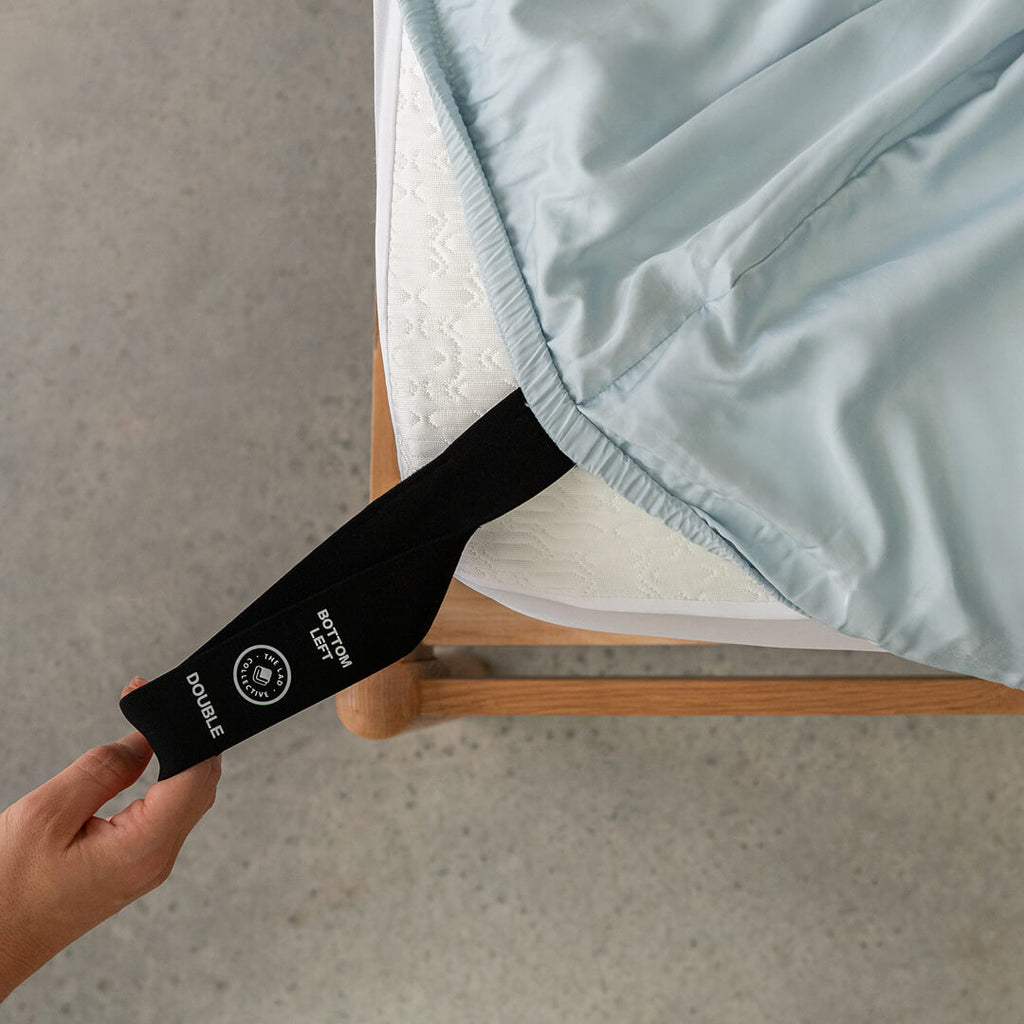 Person holding a black strap on a light blue fitted sheet, pulling it over the mattress.