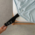 Person holding a black strap on a light blue fitted sheet, pulling it over the mattress.