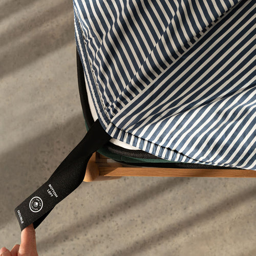 Navy and white pinstripe fitted sheet showcasing labeled corner loop strap being pulled by a hand onto the bed.