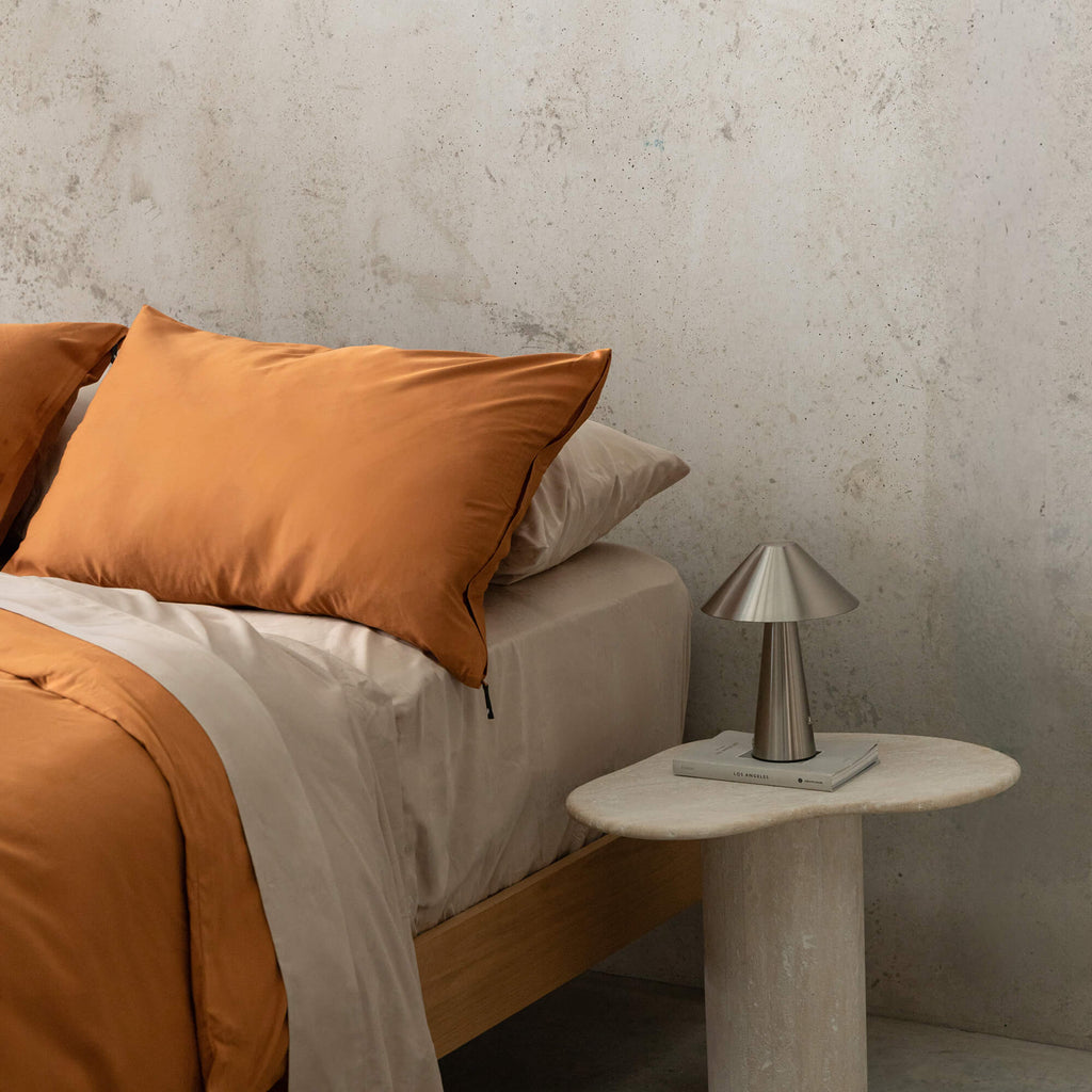 Close up of burnt orange duvet cover set on a bed, against a neutral concrete look backdrop, with a marble bedside table and lamp on the side.