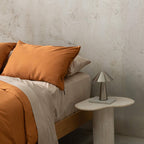 Close up of burnt orange duvet cover set on a bed, against a neutral concrete look backdrop, with a marble bedside table and lamp on the side.