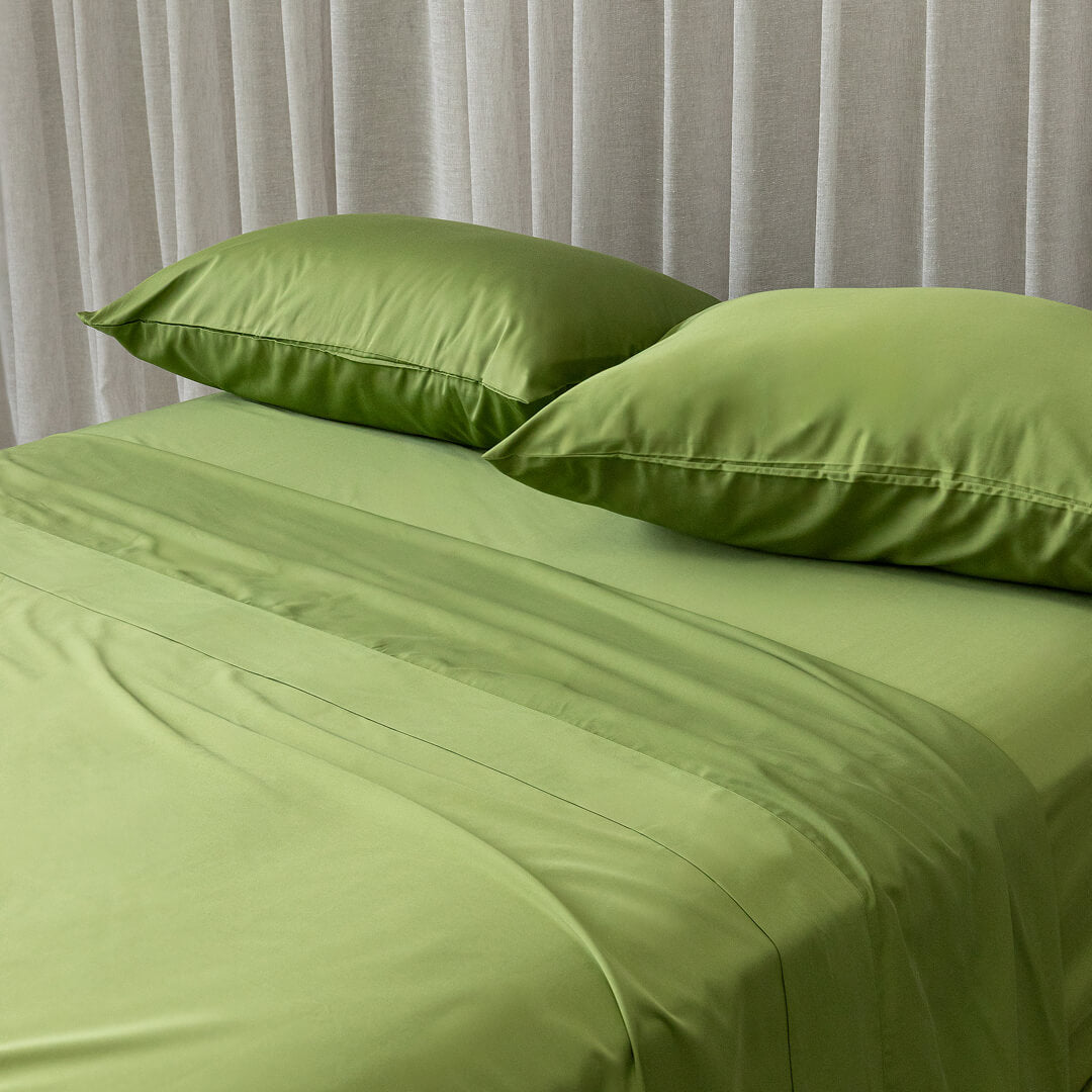 Green bed sheets and pillows on a bed with a neutral curtain background