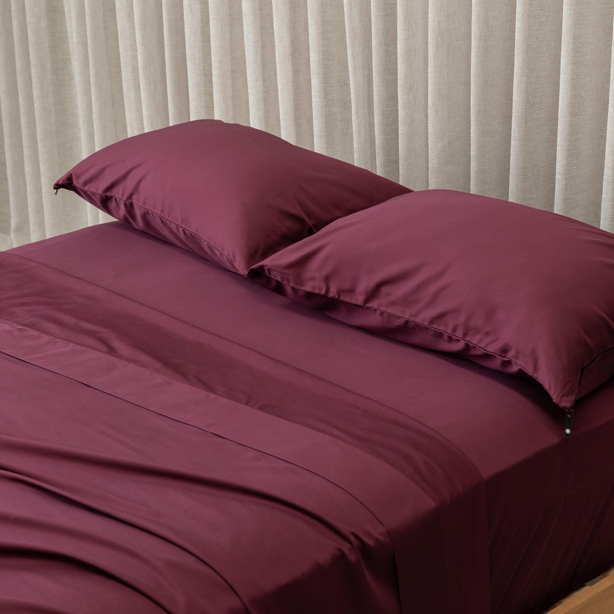 Burgundy bed sheets and pillows on a bed with light curtains in the background