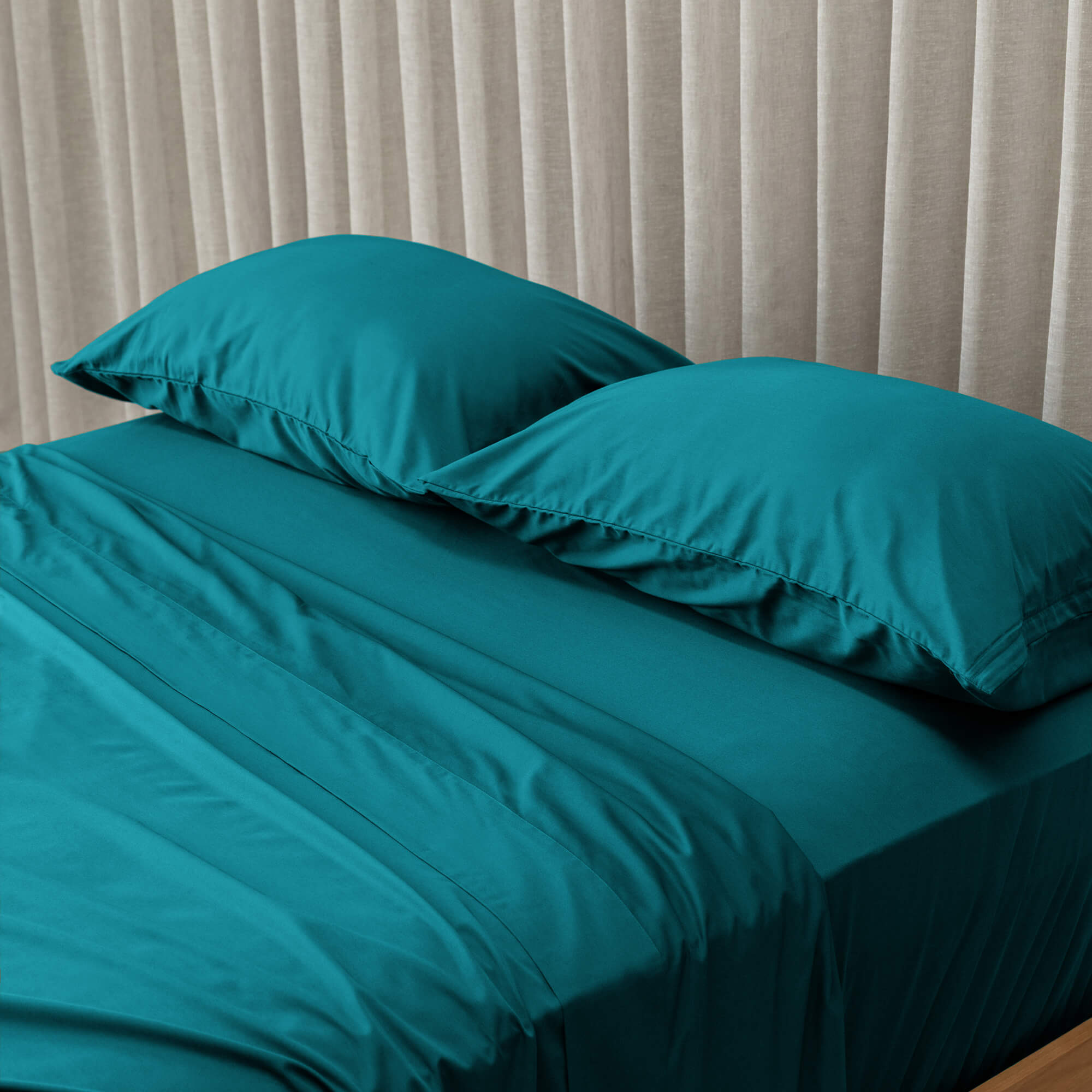 Teal bed sheets and pillows on a bed with beige curtains in the background
