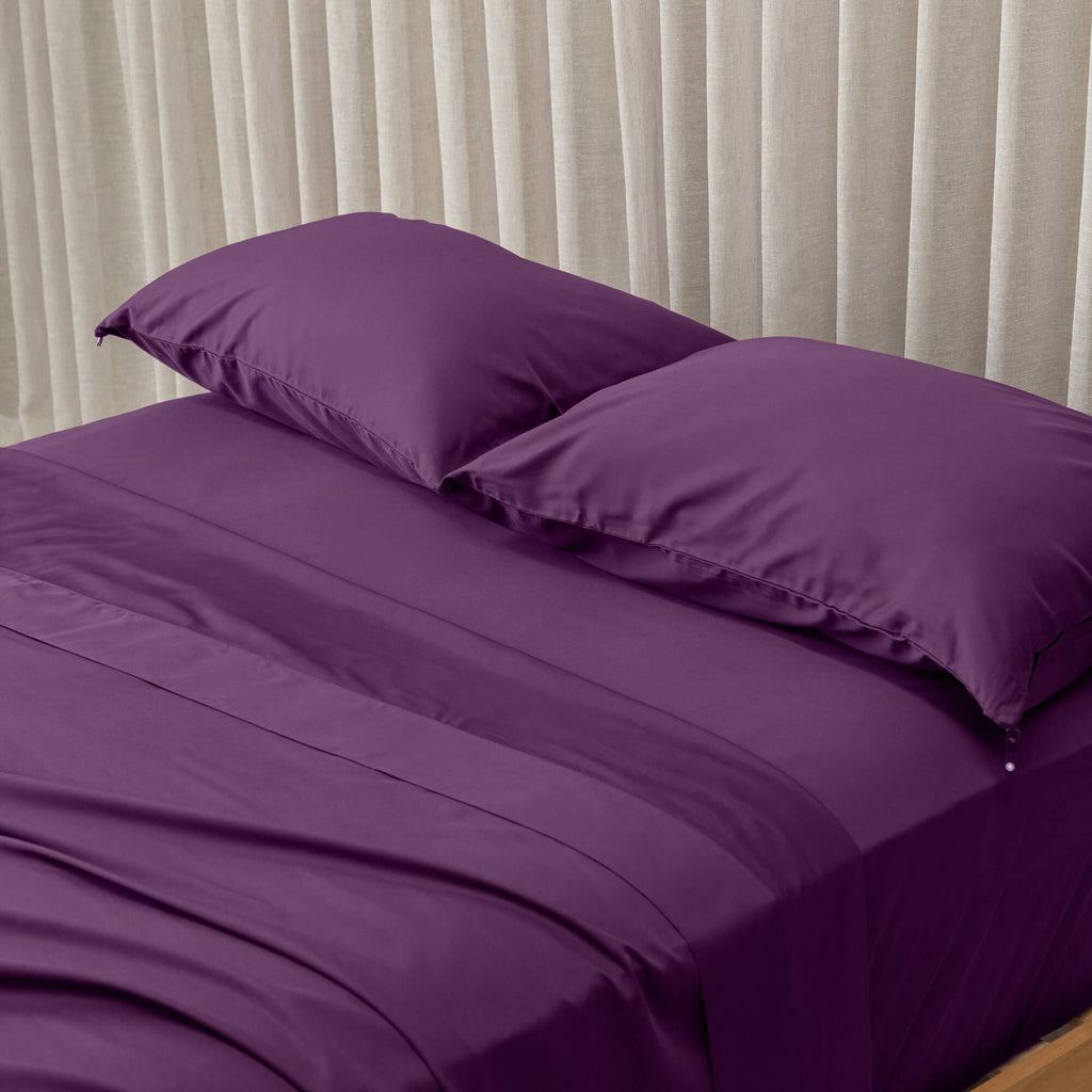 Purple bed sheets and pillows on a bed with beige curtains in the background.