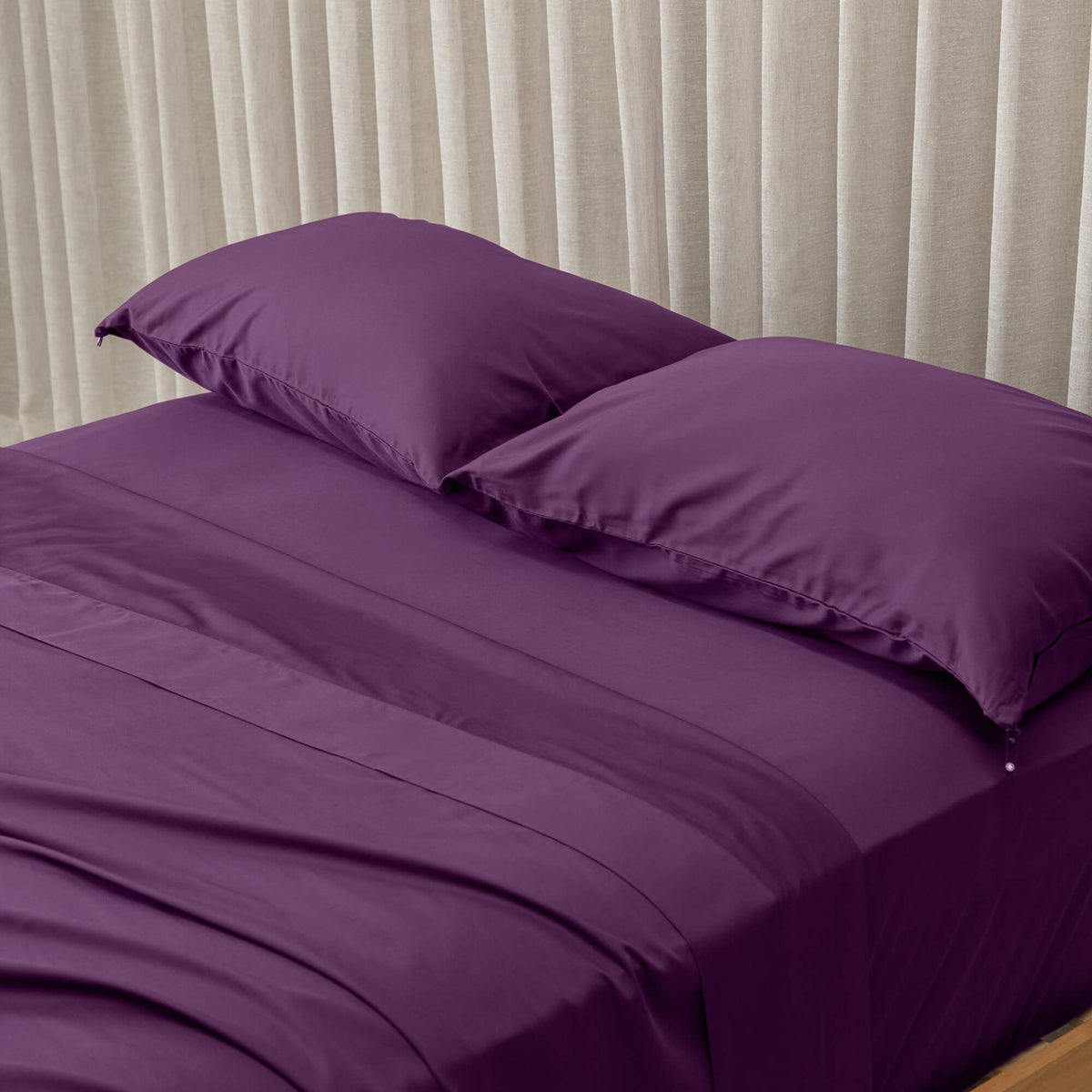 Purple bed sheets and pillows on a bed with beige curtains in the background.