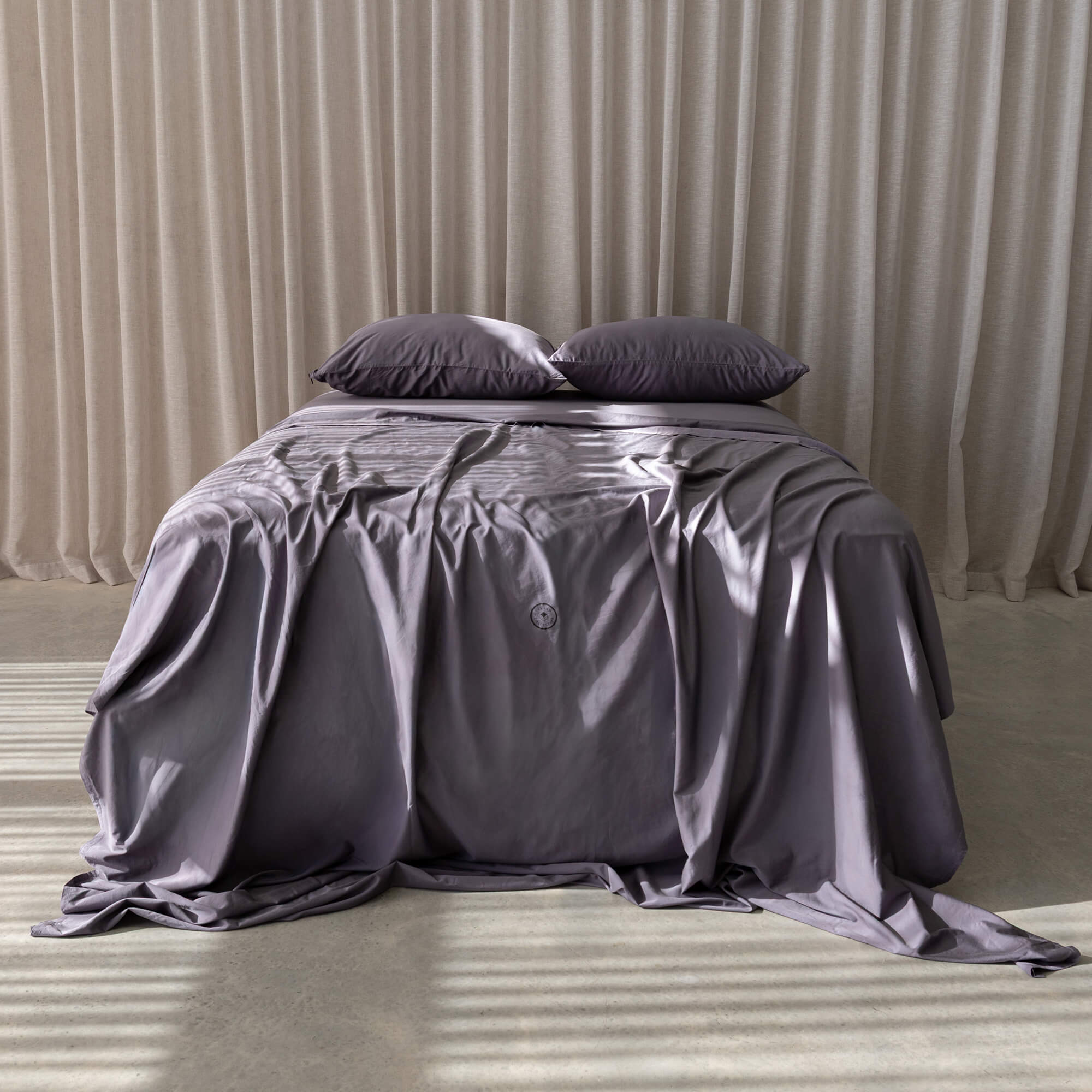 Gray bedspread and pillows on a bed with beige curtains in the background