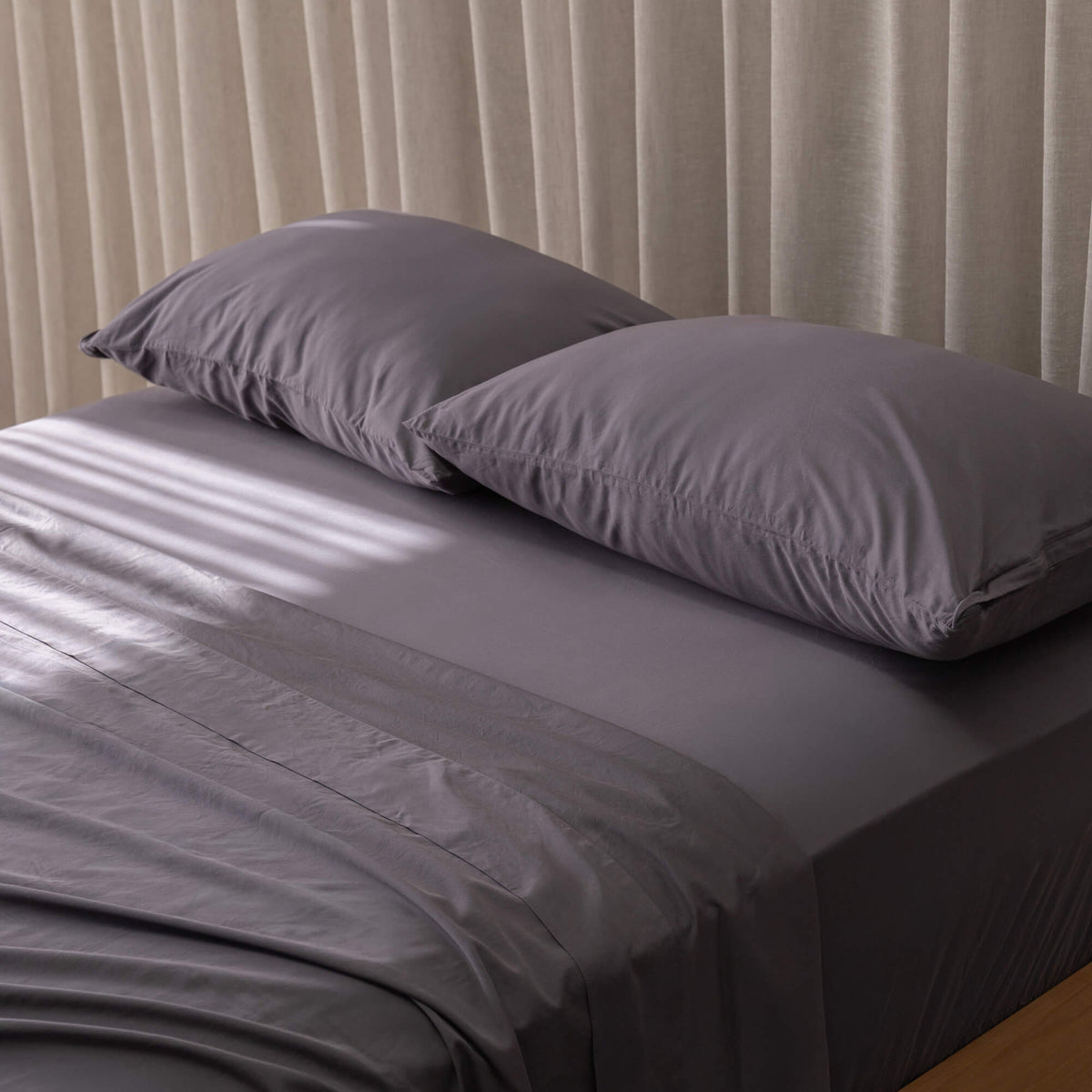 Gray bed sheets and pillows on a bed with beige curtains in the background