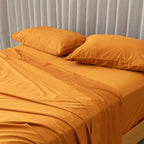 Orange bedspread and pillows on a bed with beige curtains in the background.