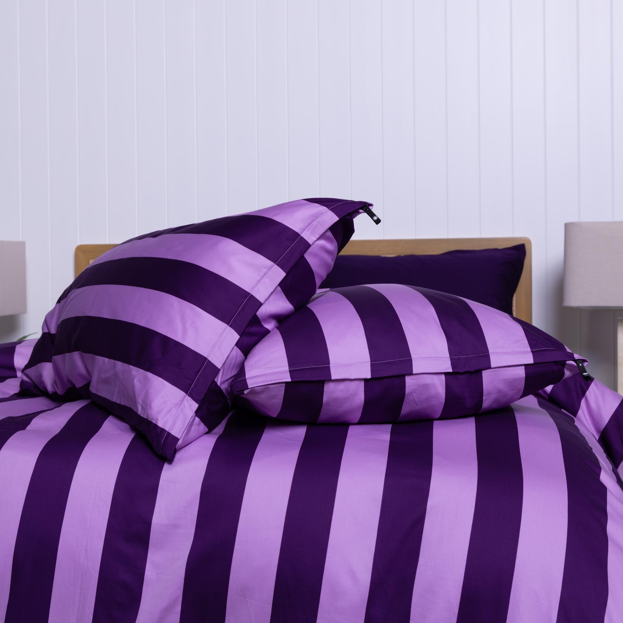 Purple and lilac striped bedding set on a bed with a white wall background and pillows on the bed.