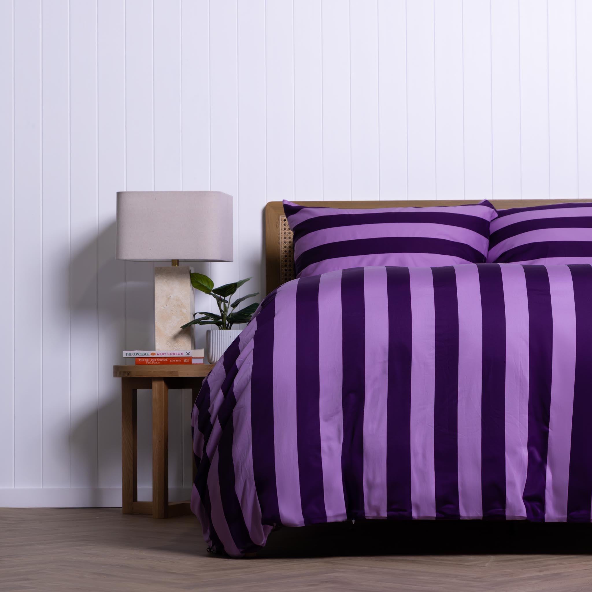 Bed with purple striped bedding next to a nightstand with a lamp and books against a white paneled wall.