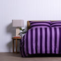 Bed with purple striped bedding next to a nightstand with a lamp and books against a white paneled wall.