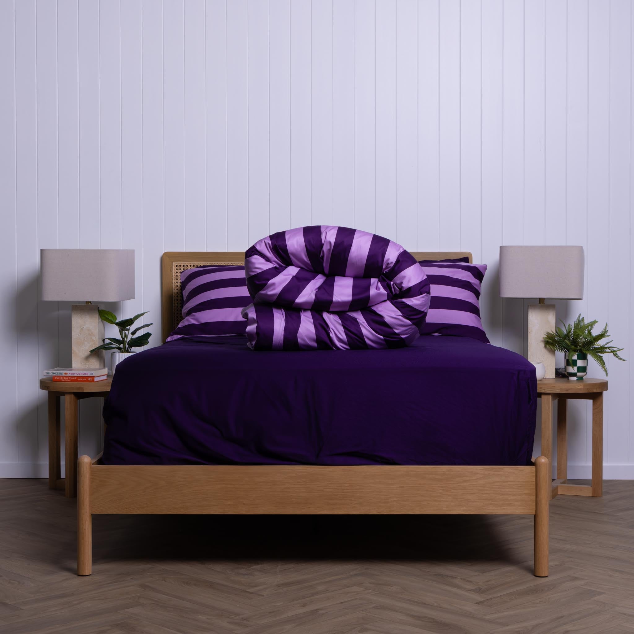 Bed with purple bedding and striped duvet cover rolled up on the bed with two lamps and plants on nightstands.