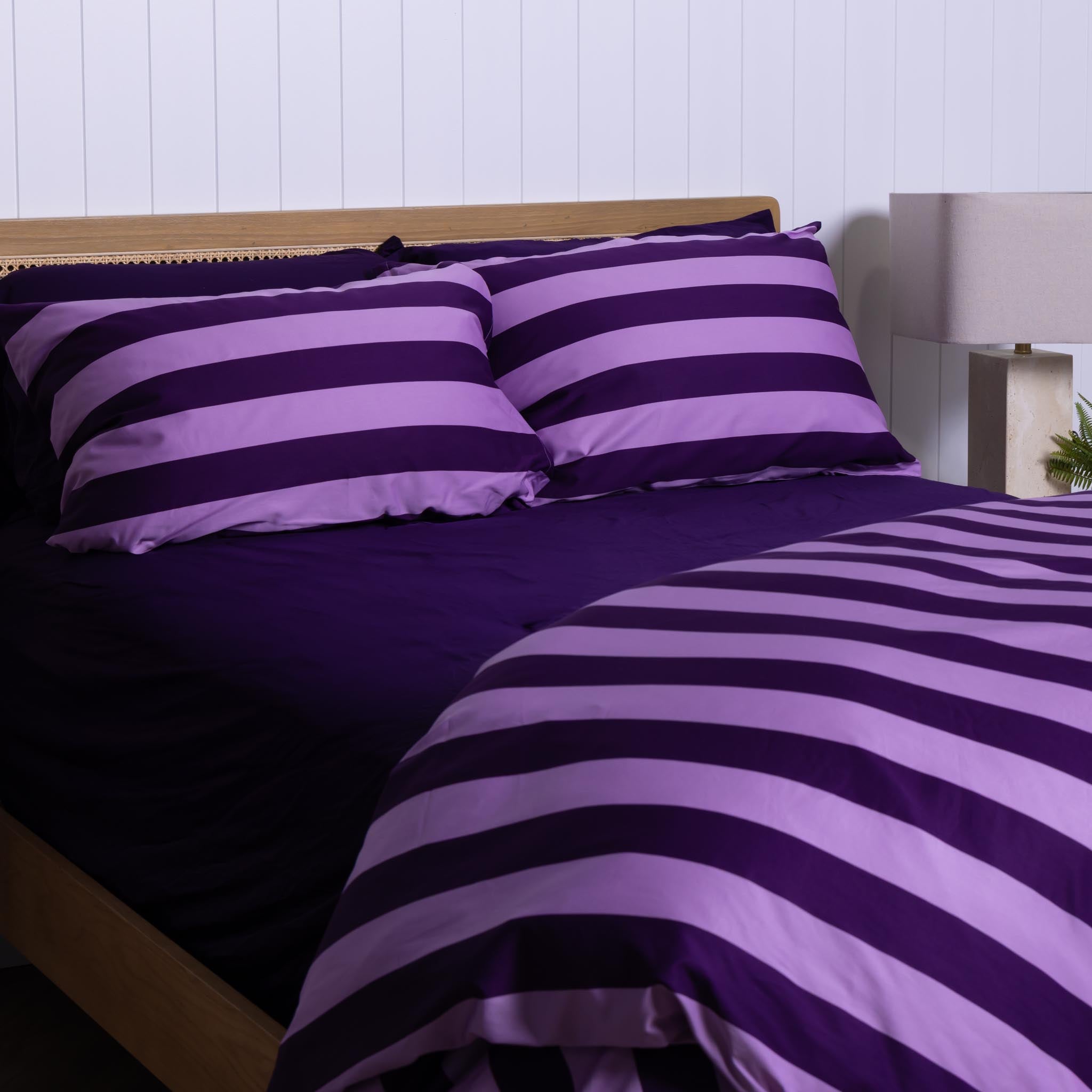 Lilac and dark purple striped bedding set on a bed with a wooden headboard - shown on an angle.