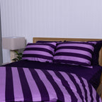 Purple and lilac striped bedding set on a bed with a lamp and plant in the background.