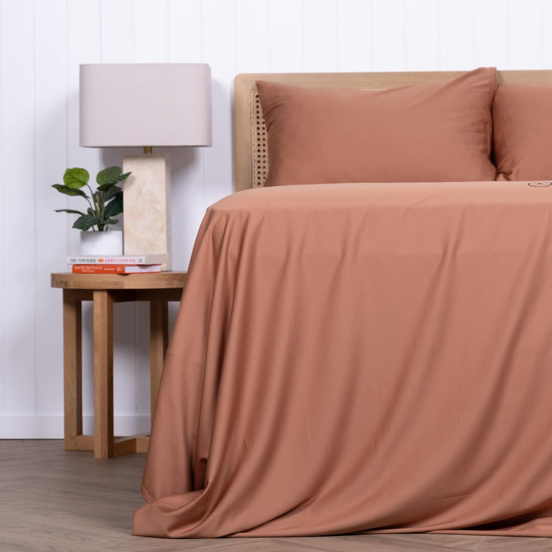 Side view of a bed styled with rust-coloured sheets and pillows next to a wooden bedside table.