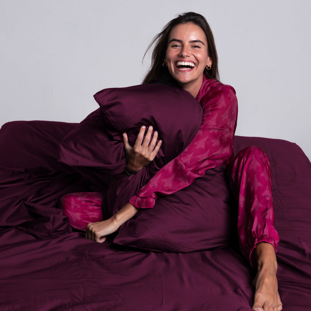 Woman laughing and hugging burgundy pillow on TLC 3.0 Bedding Set, conveying softness and joy.