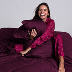 Woman laughing and hugging burgundy pillow on TLC 3.0 Bedding Set, conveying softness and joy.