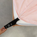 Black labelled corner loop strap on pink fitted sheet being pulled onto mattress by a hand.