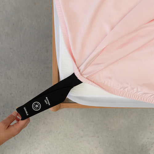 Black labelled corner loop strap on pink fitted sheet being pulled onto mattress by a hand.