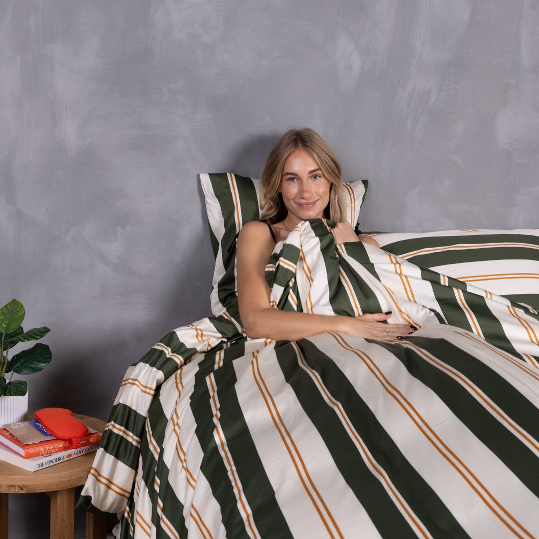 Woman sitting on a bed with green and white striped bedding against a gray wall.