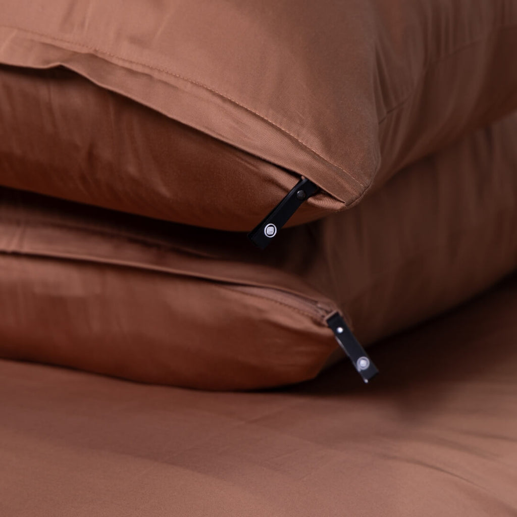 Two stacked rust-coloured zippered pillowcases with pillows inside from The Lad Collective.