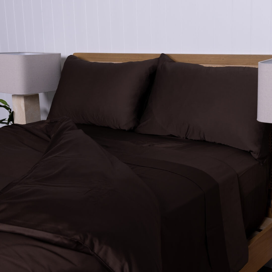 Brown bed sheets and pillows on a bed with a white wall and lamp in the background.