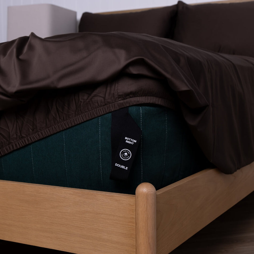 Brown fitted sheet on a wooden bed with a visible tag.