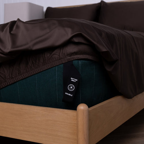 Brown fitted sheet on a wooden bed with a visible tag.